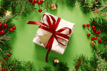 Stylish Christmas gift flat lay. Modern christmas present with red ribbon and green fir branches, red berries and gold ornaments on green background. Xmas background. Season's greeting card