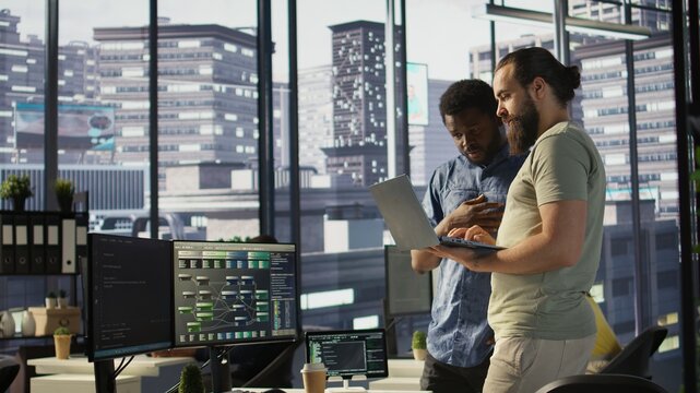 IT employees in office working together using laptop, testing and deploying programs and systems. Developer and colleague checking code on notebook, doing brainstorming in workspace, camera B