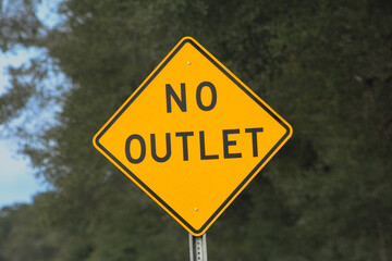 A bright yellow "No Outlet" road sign stands prominently against a blurred natural background. Sign warns drivers of a dead-end or limited exit route ahead, symbolizing caution or restriction.
