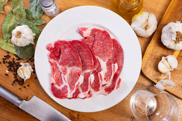 Fresh marbled pork loin head slices streaked with fat, prepared for cooking on plate, surrounded by garlic, bay leaves, and pepper for enhanced flavor on wooden table..
