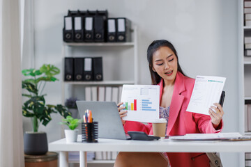 Asian business woman shows documents with clipboard graphs holds in her hand, looks at the camera loptop, smiles. Sitting at a desk with a laptop in a modern office.
