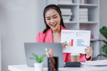 Asian business woman shows documents with clipboard graphs holds in her hand, looks at the camera loptop, smiles. Sitting at a desk with a laptop in a modern office.
