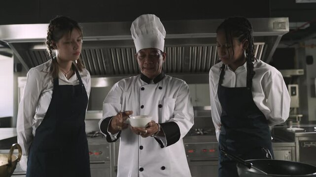 Professional Asian senior male chef in uniform with chef hat teaching cooking methods to female multiracial students listening intently and are ready to learn practical actions in restaurant kitchen. 