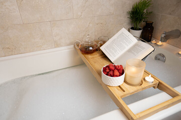 Wooden tray on a bathtub with book, candle and herbal tea. Relaxation spa time. Time for yourself.