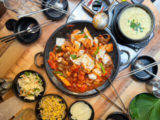 A vibrant Korean meal with jjukkumi (baby octopus) stir-fried in spicy sauce, surrounded by fresh vegetables, rice cakes, and various side dishes on a wooden table. Authentic Korean dining experience.