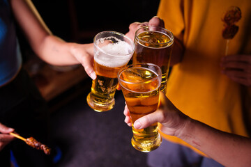 Close-up of friends clinking glasses of beer, celebrating together at a bar.The lively atmosphere captures laughter, camaraderie,good times, as young friends enjoy a night out with refreshing drinks