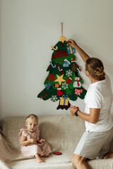 Father with little daughter sit on the sofa and put on toys on felt christmas tree during the holiday season. A family enjoying a cozy Christmas evening at home