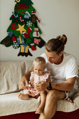 Father with little daughter sit on the sofa and put on toys on felt christmas tree during the holiday season. A family enjoying a cozy Christmas evening at home