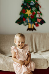 Cute smiling little girl waiting for vhristmas, sit at home on the sofa near just decorated felt christmas tree, look at the camera and smiling