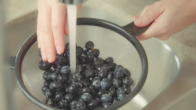 Woman's hands washing blueberries in a strainer basket under tap water. Slow motion. 
