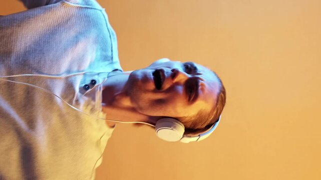 Vertical video. Emotional guy. Dance moment. Positive creative young man listening music with headphones while moving isolated over yellow wall background.