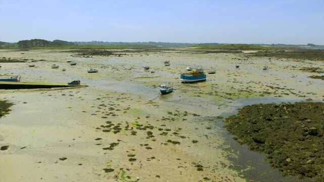 Small fishing boats sit on Brittany&rsquo;s sandy flats during low tide, revealing seaweed-covered rocks and a rugged coastal landscape under clear skies, creating a vibrant maritime scene.