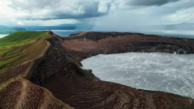 Aerial close look inside the crater of Taal Volcano in the Philippines, lush greenery climbing the crater walls. Geological formations add texture, with Taal Lake visible in the rainy distance.