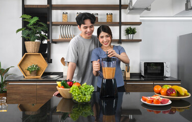 Young asian woman using blending machine mixing fruit and vegetable in to smoothie. Her boyfriend stand smiling behind. The kitchen counter full of various kinds of fruit and vegetable.