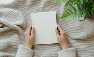 Fototapeta premium Person holding a blank notebook while wrapped in a cozy sweater, with a soft blanket and a green plant nearby.
