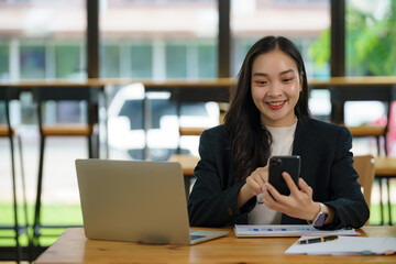 A woman sits at a desk with a laptop and a cell phone. She smiles and looks at her cell phone.