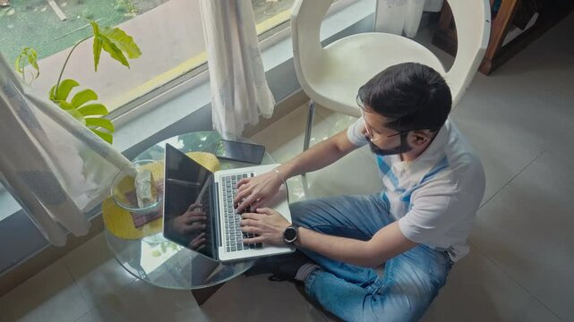 Laptop, wireless technology, internet. Top view of an Indian adult male using laptop while sitting on the floor near window side at home.