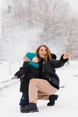 Smiling mom and little son are standing in the snow, with holding sparkler