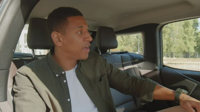 Irritated impatient black male passenger sitting on backseat of car, checking the time on wristwatch, being late to business meeting and asking taxi driver to hurry up.