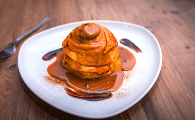 A stack of fluffy pancakes drizzled with caramel sauce, served on a white plate. The rich caramel flows down, creating a mouth-watering dessert presentation on a wooden table.