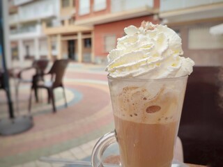 A glass of coffee with whipped cream served in an outdoor cafe, featuring a blurred urban background