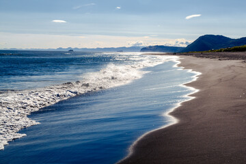 View from the Khalaktyrsky beach to the Pacific Ocean against the background of the blue sky. On the horizon of the black sand beach, cliffs and ocean waves are visible. Kamchatka Peninsula, Russia.