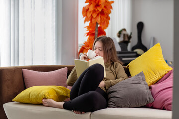 Young woman reading a book while drinking coffee on sofa at home