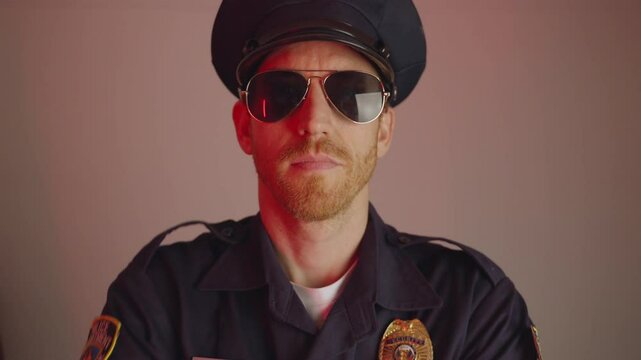 Tough looking cop or police officer in official uniform looks at camera and then takes off his sunglasses and winks. Serious and handsome policeman on solid background. Funny quirky medium shot