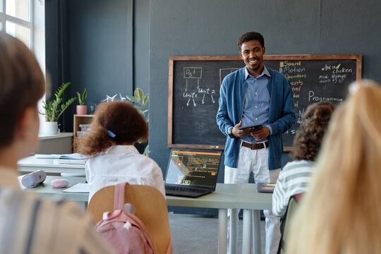 Wide angle view of school classroom with group of children and smiling African American man teaching IT copy space