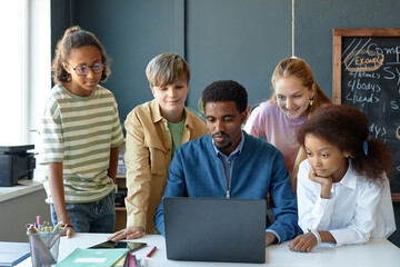 Front view portrait of young male teacher with diverse group of children using computer together in...
