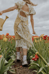 A girl in a yellow dress and apron with a water can in a red and green tulip flower field on a gloomy rainy cloudy sky background happily running around. 