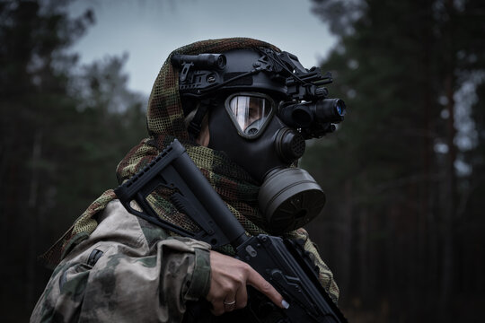 A girl soldier in a gas mask and a tactical helmet with a night vision monocular in the forest at dusk.