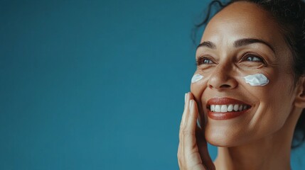 A 50 years old woman smiles and applies cream on her face against a blue background