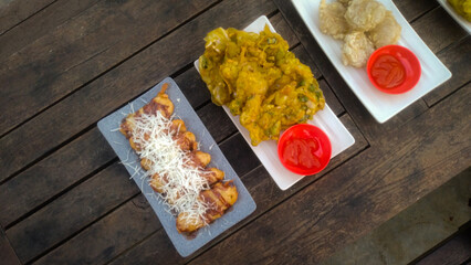 A top-down view of an assortment of Indonesian street food served on a rustic wooden table. A fried banana with grated cheese on top, a plate of vegetable fritters.