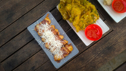 A top-down view of an assortment of Indonesian street food served on a rustic wooden table. A fried banana with grated cheese on top, a plate of vegetable fritters.