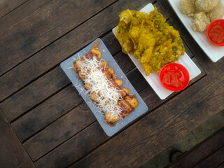 A top-down view of an assortment of Indonesian street food served on a rustic wooden table. A fried banana with grated cheese on top, a plate of vegetable fritters.