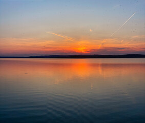 A serene lake sunset with vivid orange and blue hues reflecting on calm waters, creating a peaceful and picturesque scene against a mountain silhouette.