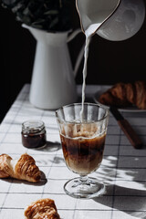Pouring milk into coffee in a transparent glass, cozy table set background. Vertical format. Latte with croissants in modern still life.