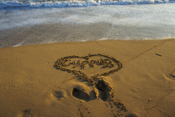 A heartfelt message drawn in the sand at the beach during sunset, surrounded by gentle waves.