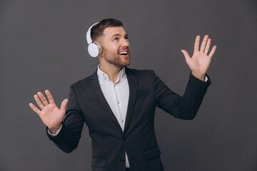 A happy smiling bearded businessman in a suit is dancing and singing attentively listening to headphones against a gray studio background