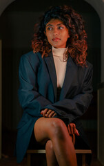 A woman in a black jacket and a white blouse is sitting on a wooden chair. She has brown hair and is wearing a necklace