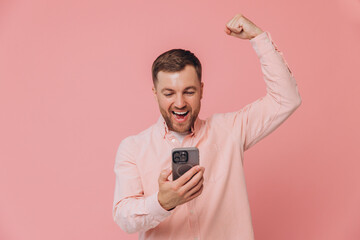 We've got a winner! Happy young man in pink shirt looking at phone screen with victory expression, isolated on pink gradient background