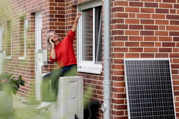 Smiling woman sitting on heat pump and talking though smart phone near window