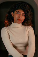 A woman with curly hair is wearing a white sweater and sitting in a room. She has a serious expression on her face