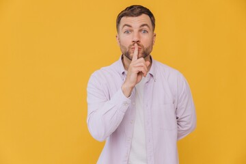 Cute unshaven man in purple shirt holding finger near mouth and showing being quiet isolated on yellow background