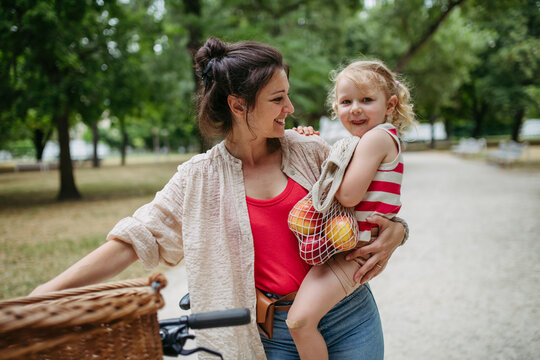 Mother carrying little girl and groceries in gthe city park pushing bicycle