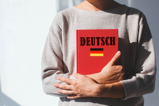 young woman in beige sweater holds red german language textbook with german inscription and german flag, foreign language learning concept. her face is not visible,