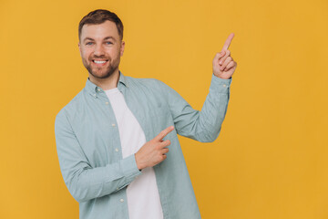Cute happy unshaven man in mint shirt showing fingers up at copy space isolated on yellow background