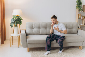 Fototapeta premium Sick man holding his chest in pain while coughing in the living room. Sore throat and cough, man with lung pain at home, health problems concept