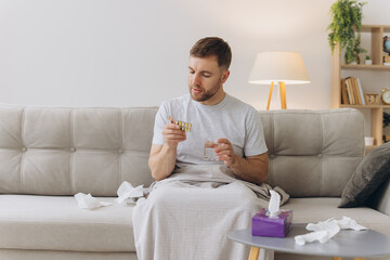 Sick man suffering from fever and headache holds blisters with pills while sitting on the sofa. Treatment and illness concept